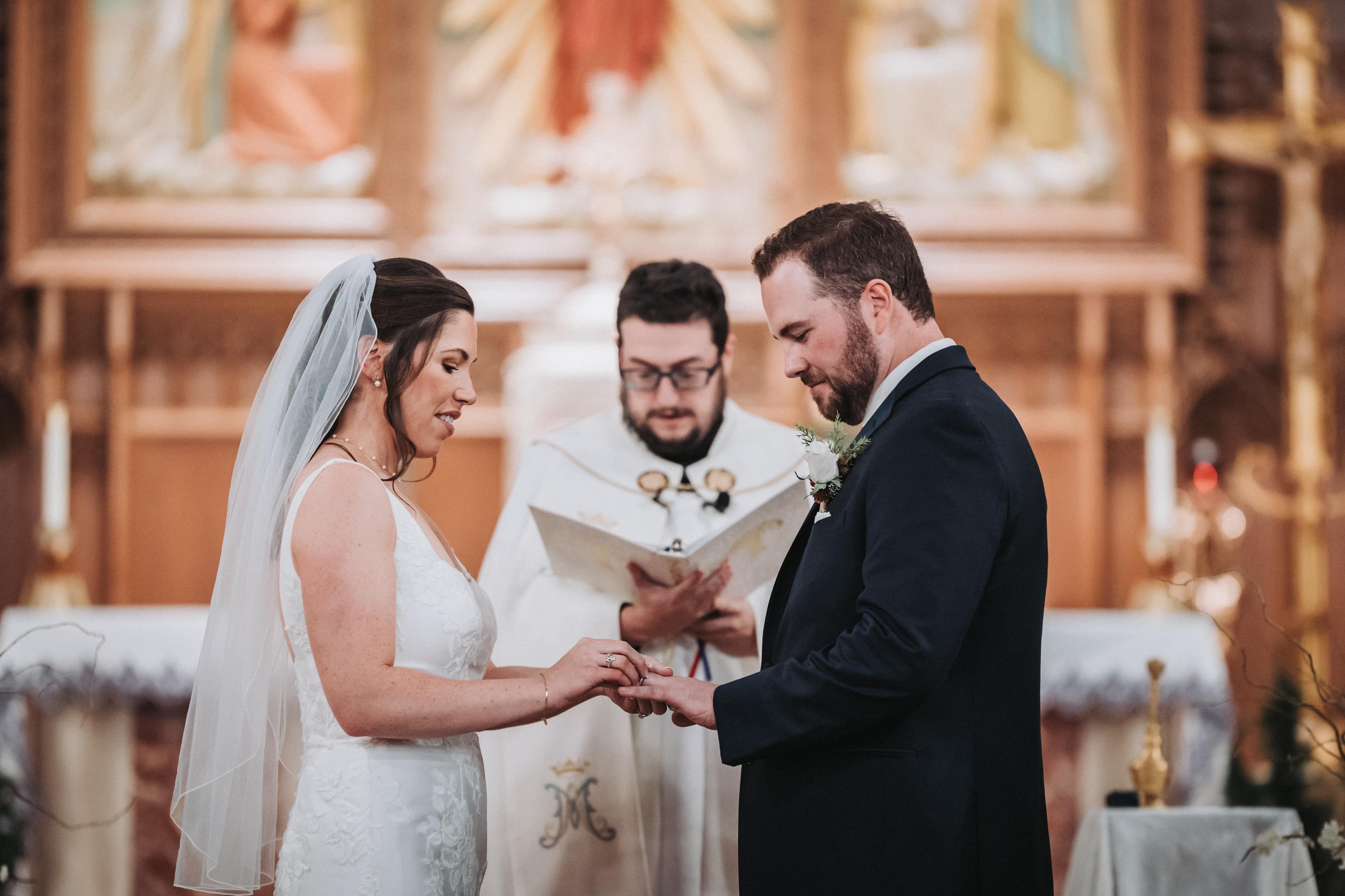 Bride and groom exchanging rings as their officiant reads from the ceremony book, a clip-on microphone visible on his vestment