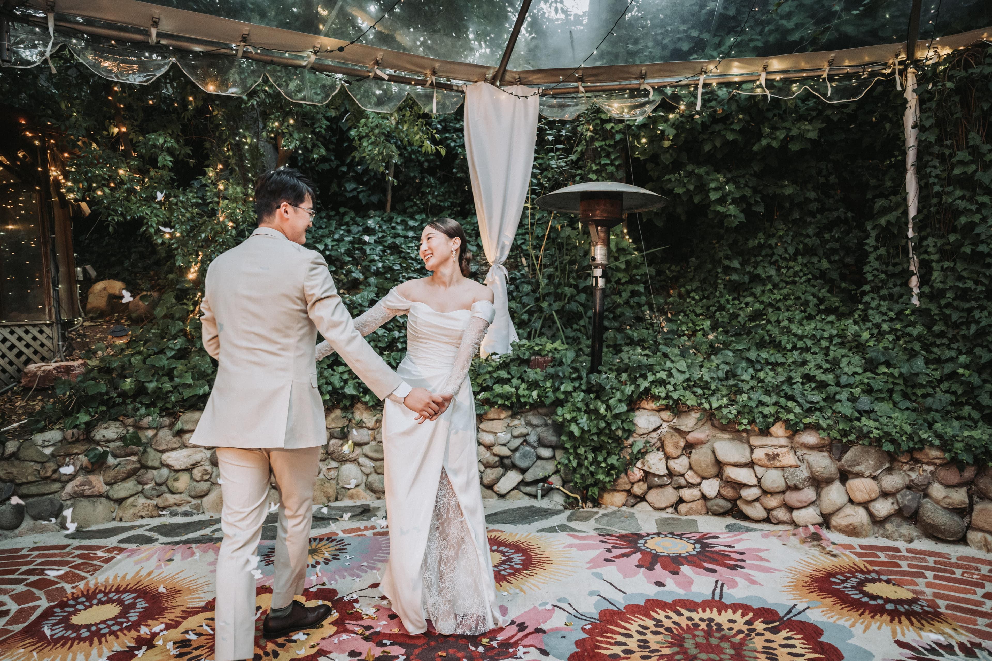 Couple holding hands and laughing on a patterned outdoor rug under a clear-top tent strung with lights, an ivy-covered wall behind them