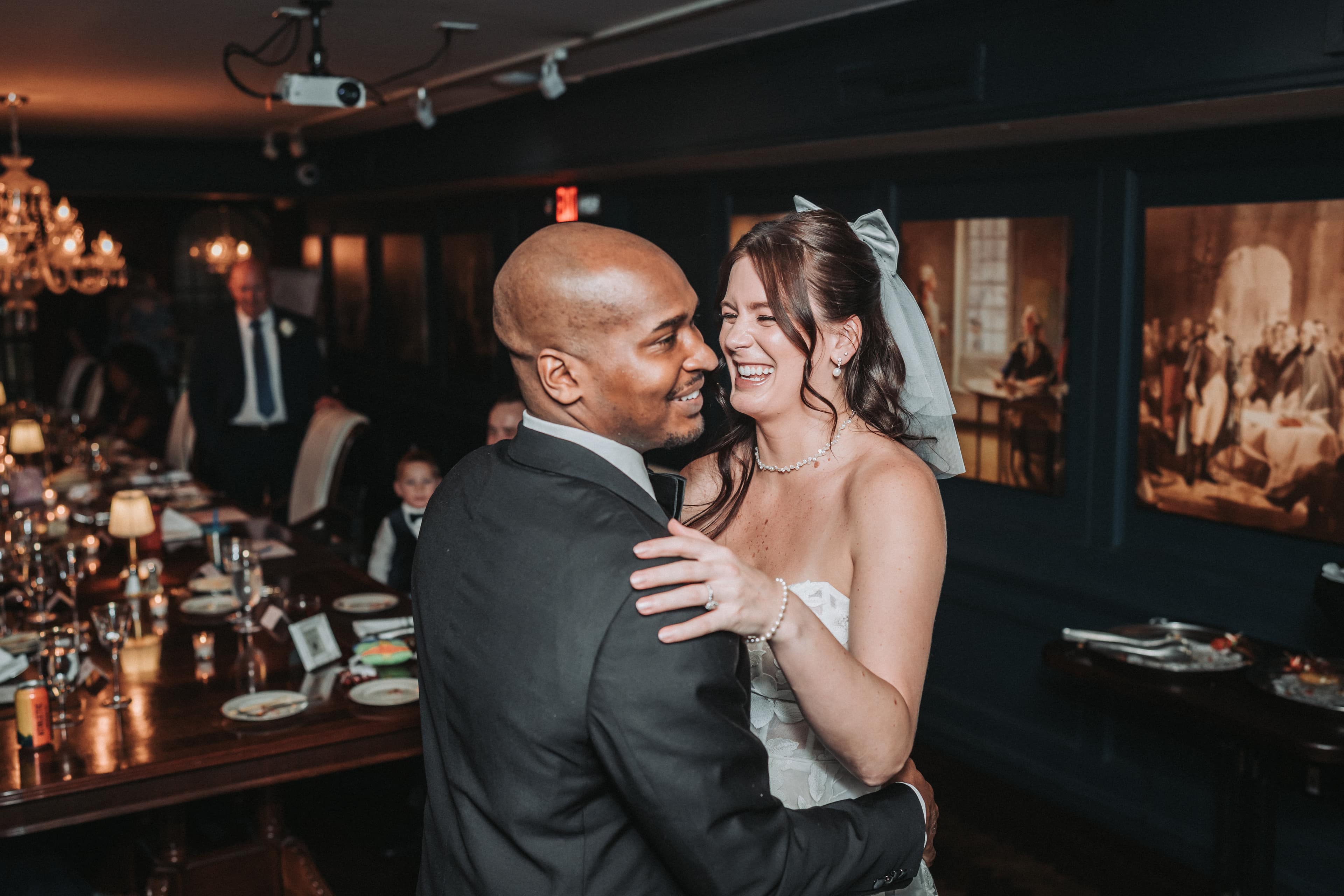 Newlyweds laughing during their first dance as guests watch from the dinner table behind them