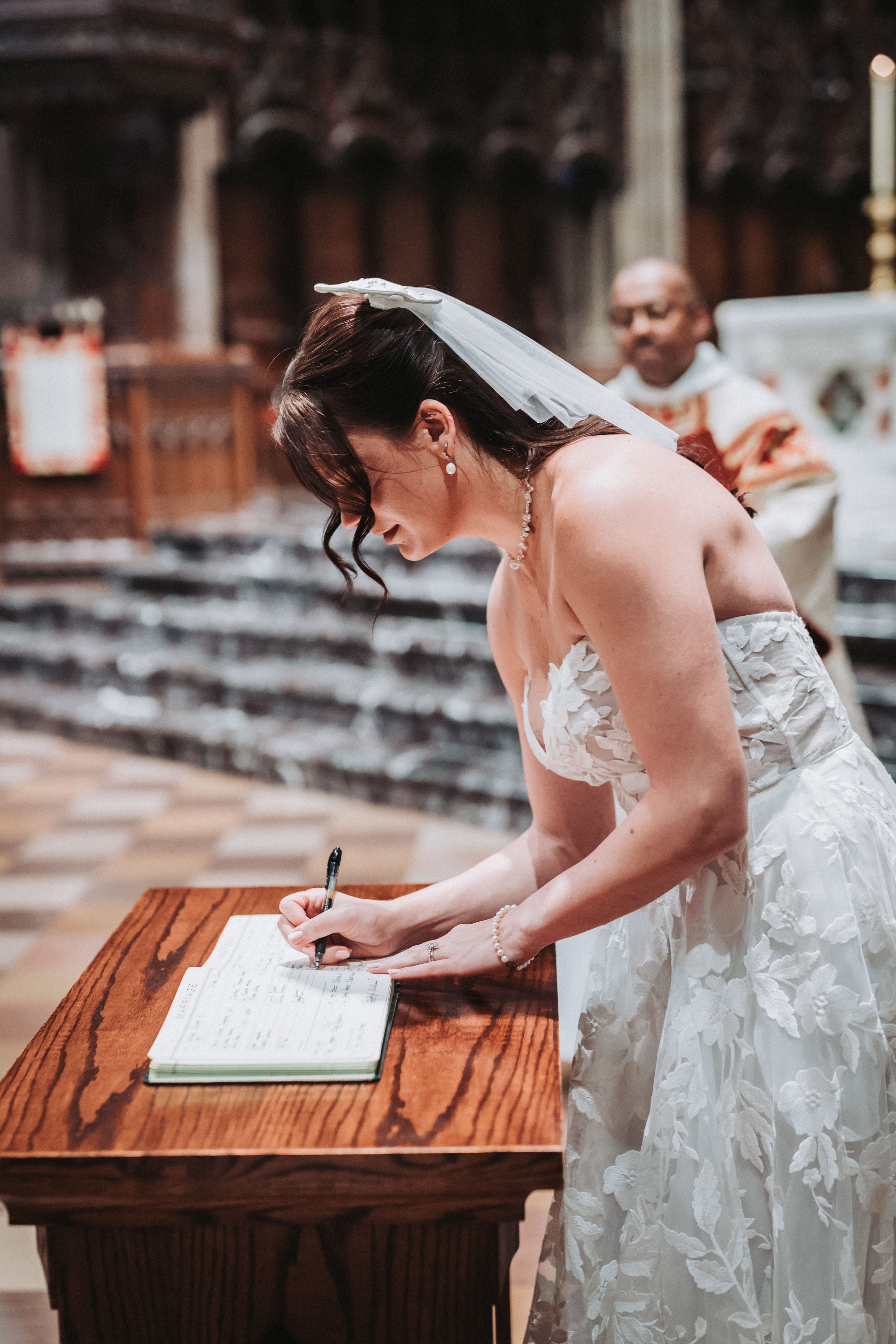 A bride in a lace strapless gown and short veil leans over a wooden lectern to sign the marriage register as the officiant looks on behind her