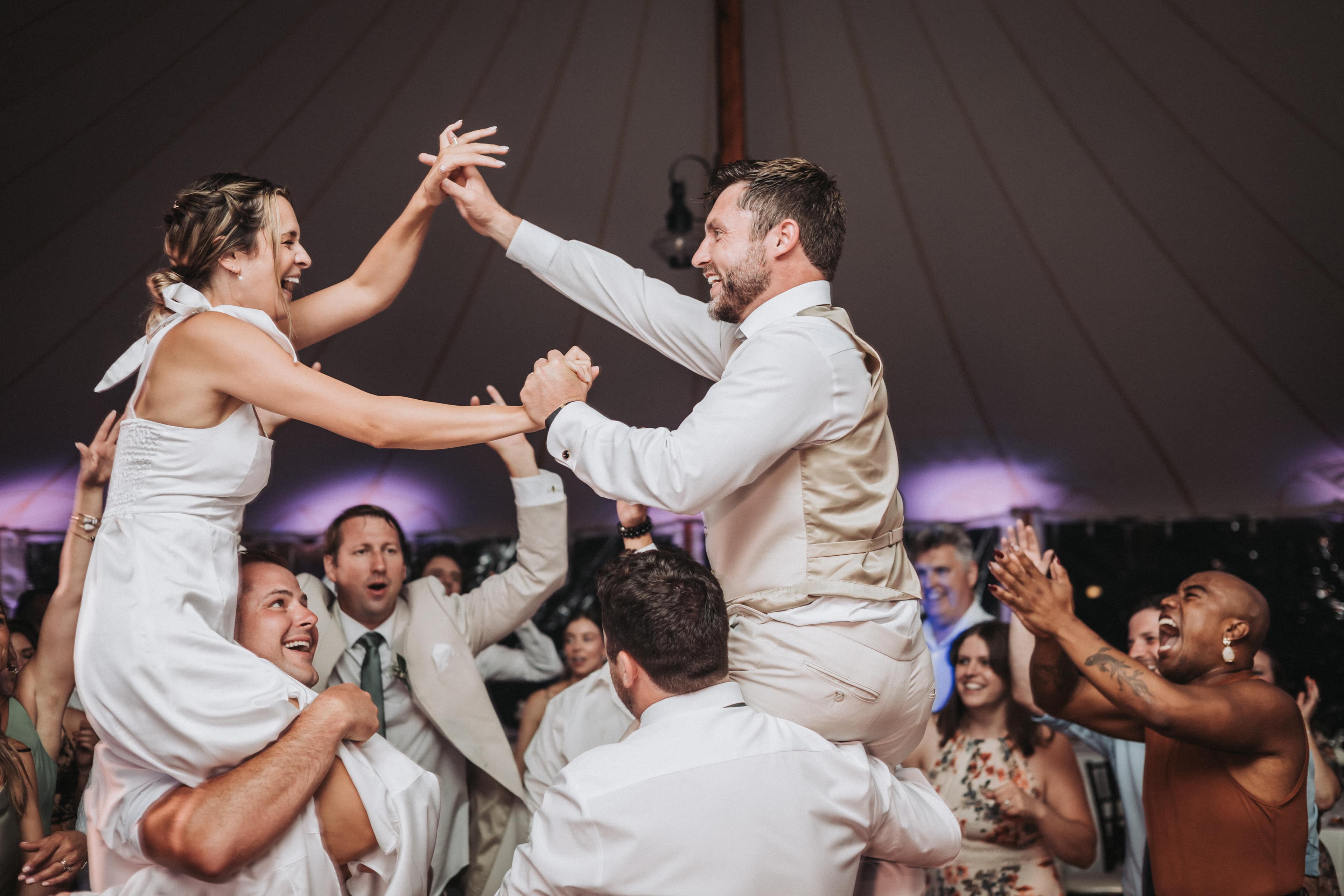 Late in the reception under a sailcloth tent, the bride in a short white dress and the groom in a tan vest are lifted onto friends' shoulders, reaching to clasp hands while guests cheer around them