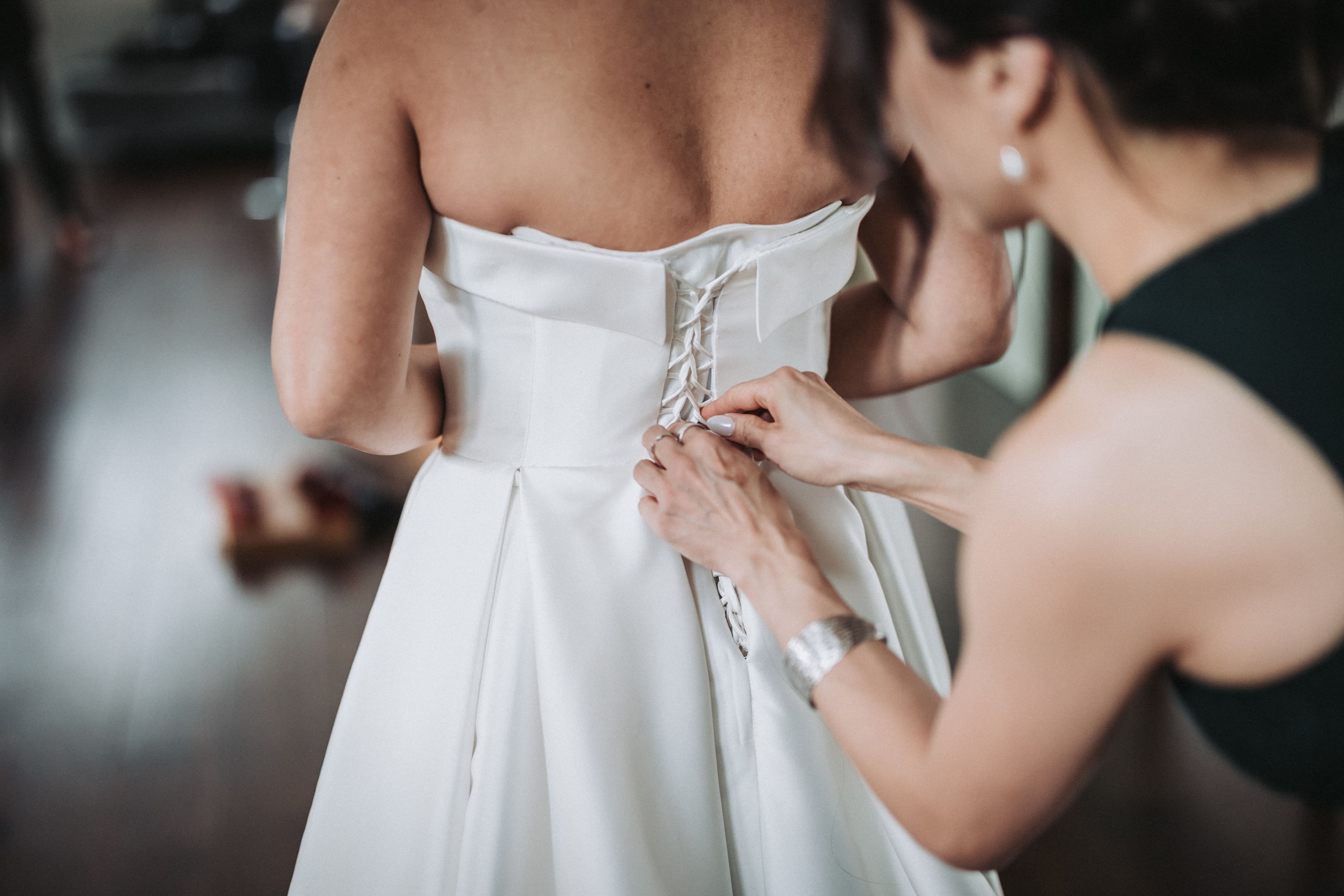 A woman in a dark sleeveless dress laces up the back of a bride's strapless ivory satin gown, her hands pulling the cord through the last loops at the waist