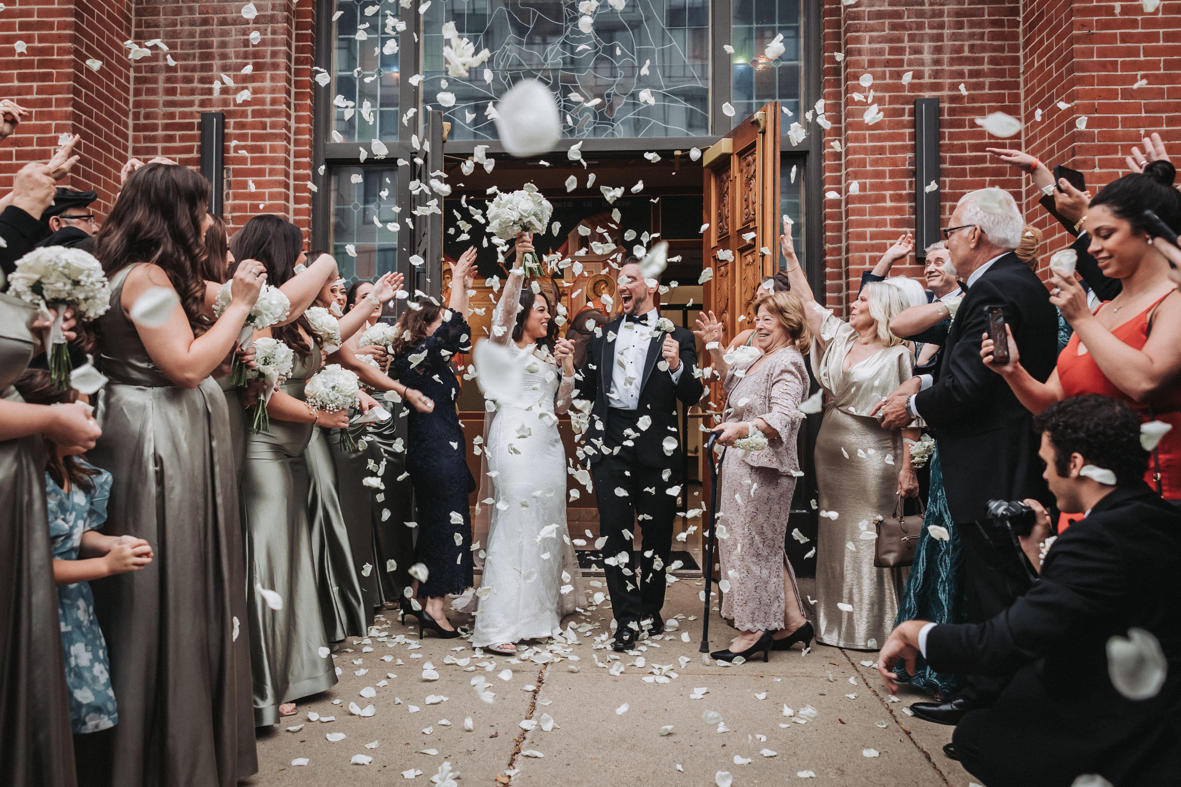 Outside a brick church, guests throw white rose petals as a laughing bride and groom walk out through its carved wooden doors, while guests on the right hold up their phones to film them