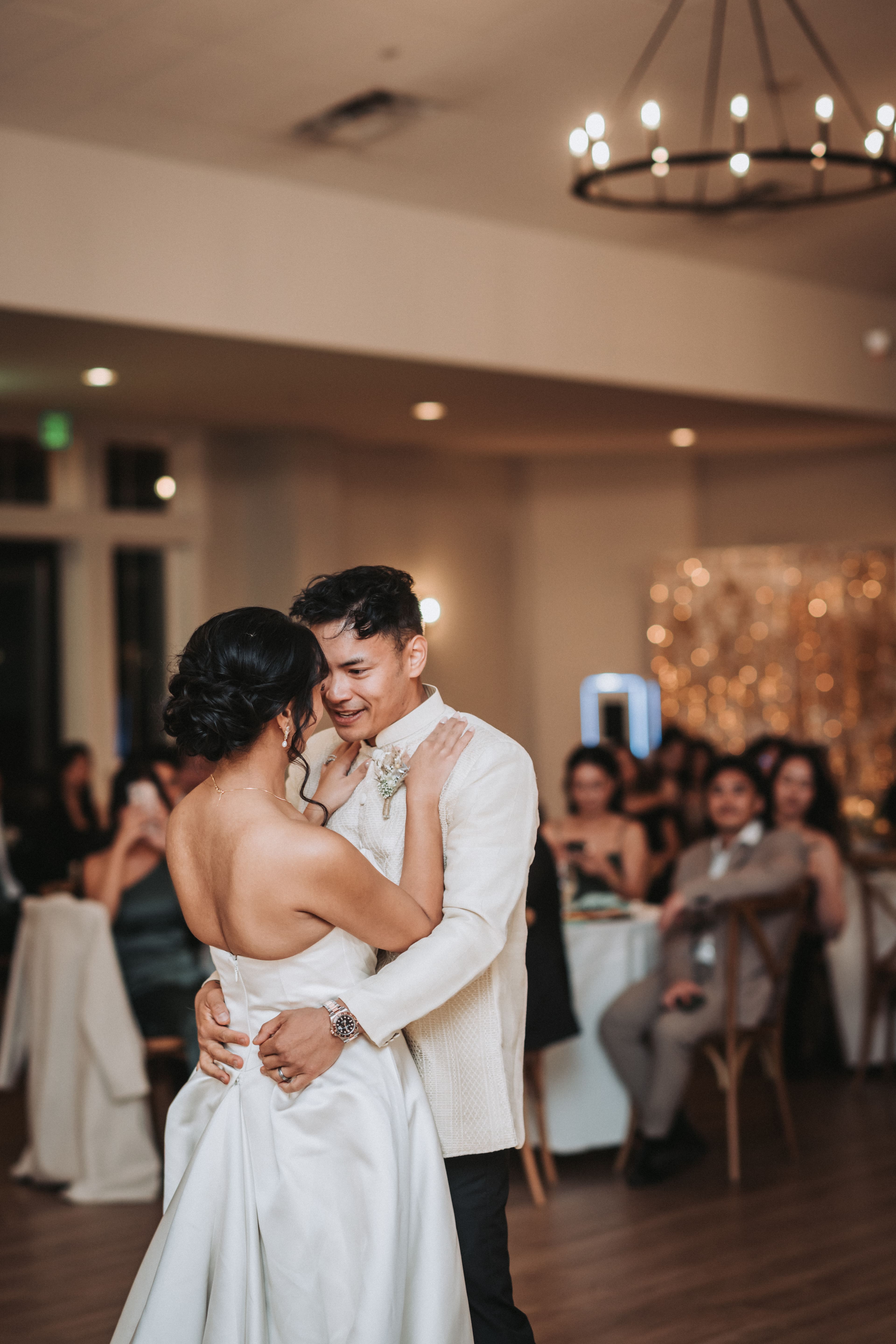 Couple slow dancing under a candle chandelier while guests watch from their tables, one of them holding up a phone to film