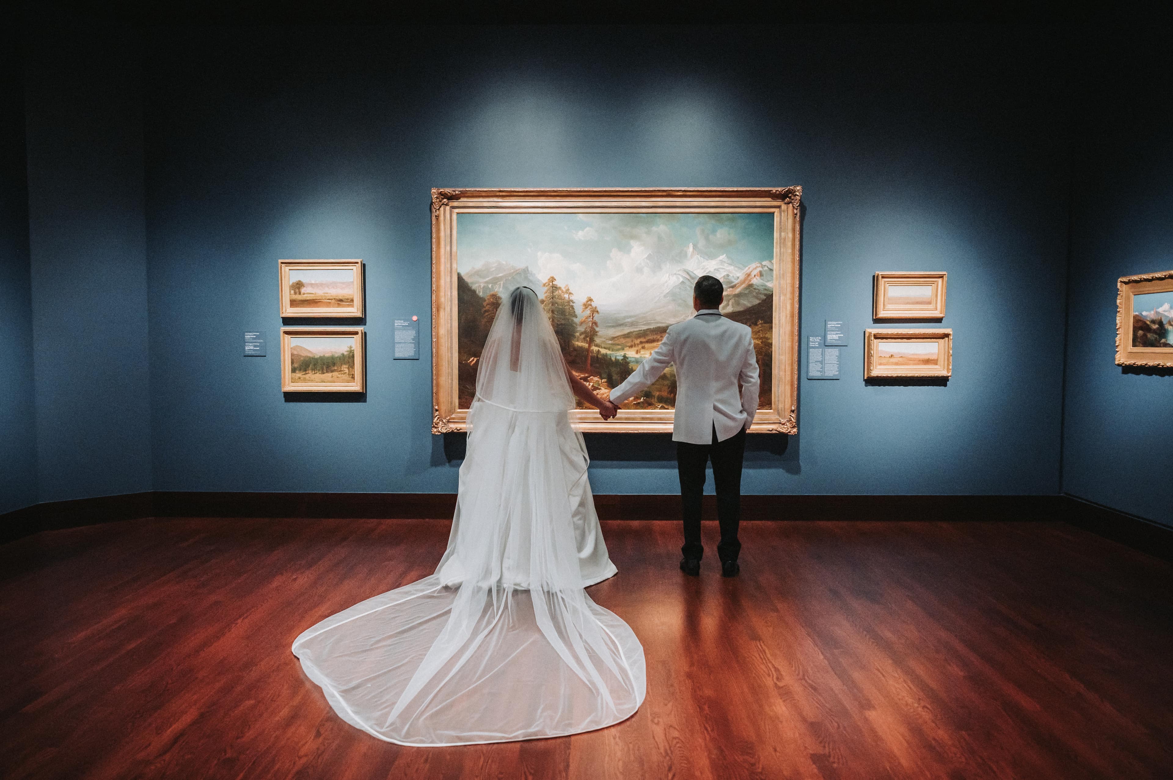 Newlyweds standing hand in hand with their backs to the camera, looking at a large framed landscape painting in a museum gallery, the bride's veil and train spread across the wood floor