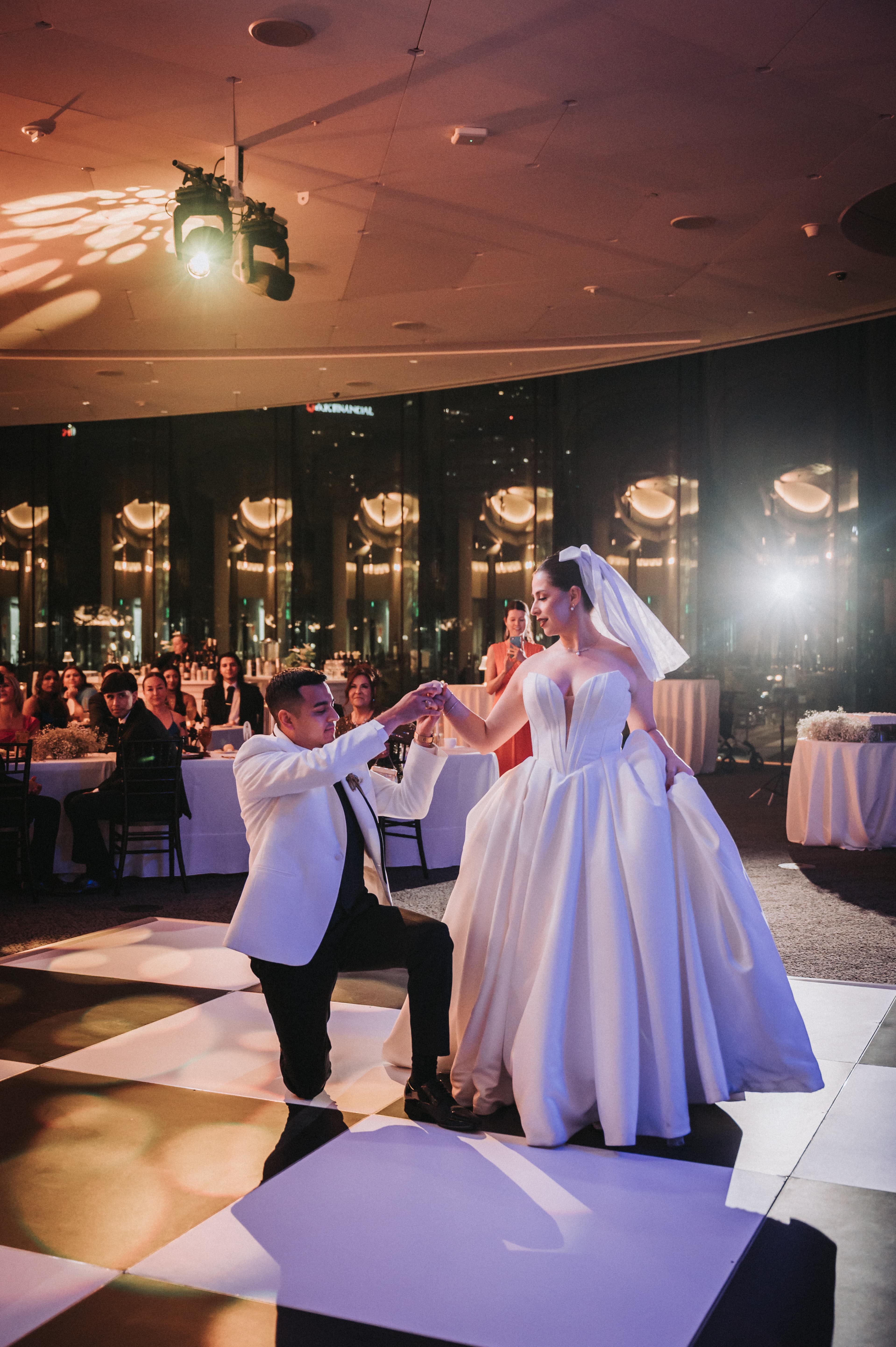 Groom kneeling to take the bride's hand on a lit dance floor during their first dance, a guest behind them filming it on her phone