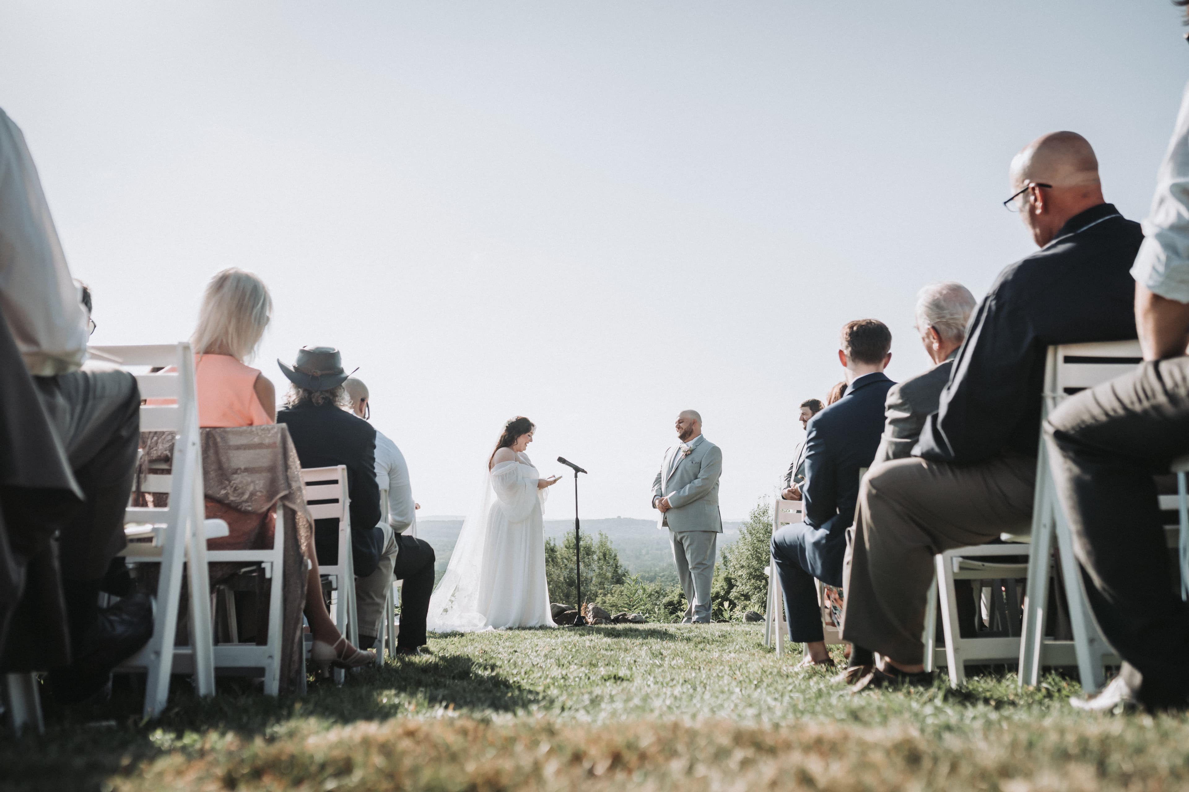 Outdoor hilltop ceremony seen from low in the aisle between rows of seated guests, the bride reading her vows at a microphone stand while the groom listens