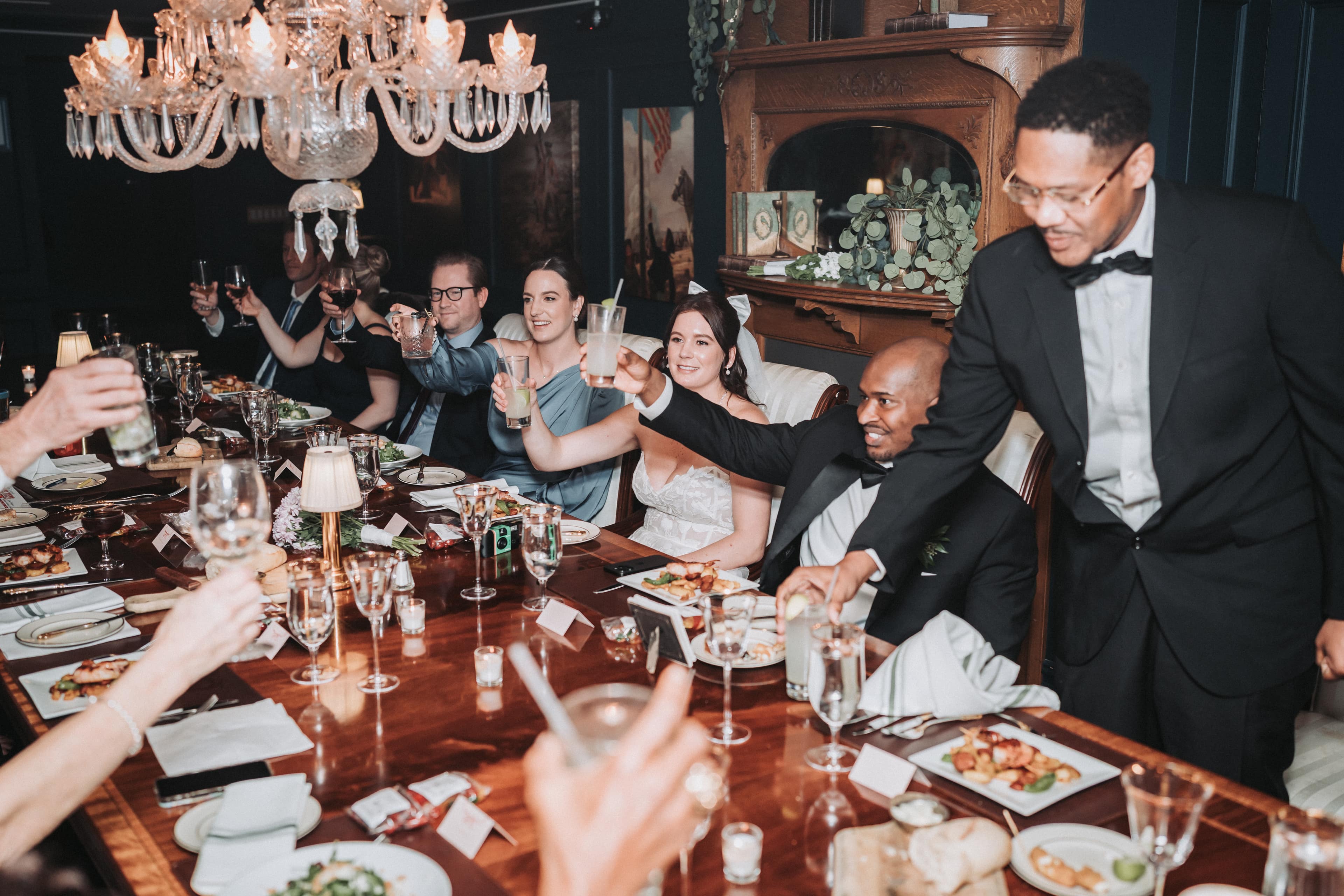 Under a crystal chandelier, a bride and groom and their guests raise their glasses in a toast around a long candlelit dinner table as a guest in a bow tie leans in to clink