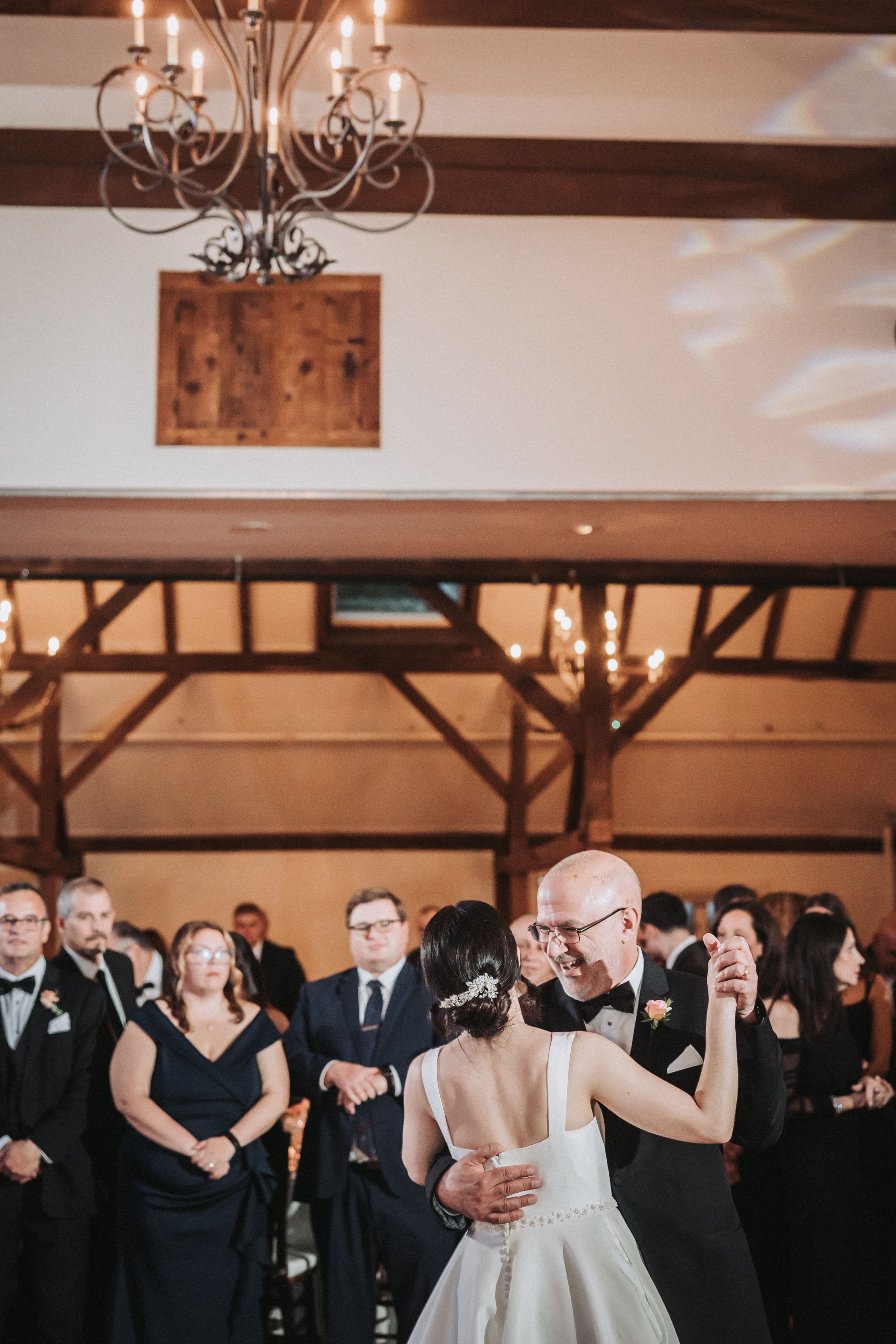 In a timber-beamed barn under a wrought-iron candle chandelier, a bride with jeweled pins in her hair dances with a grinning older man in a tuxedo while guests in black tie stand in a ring and watch