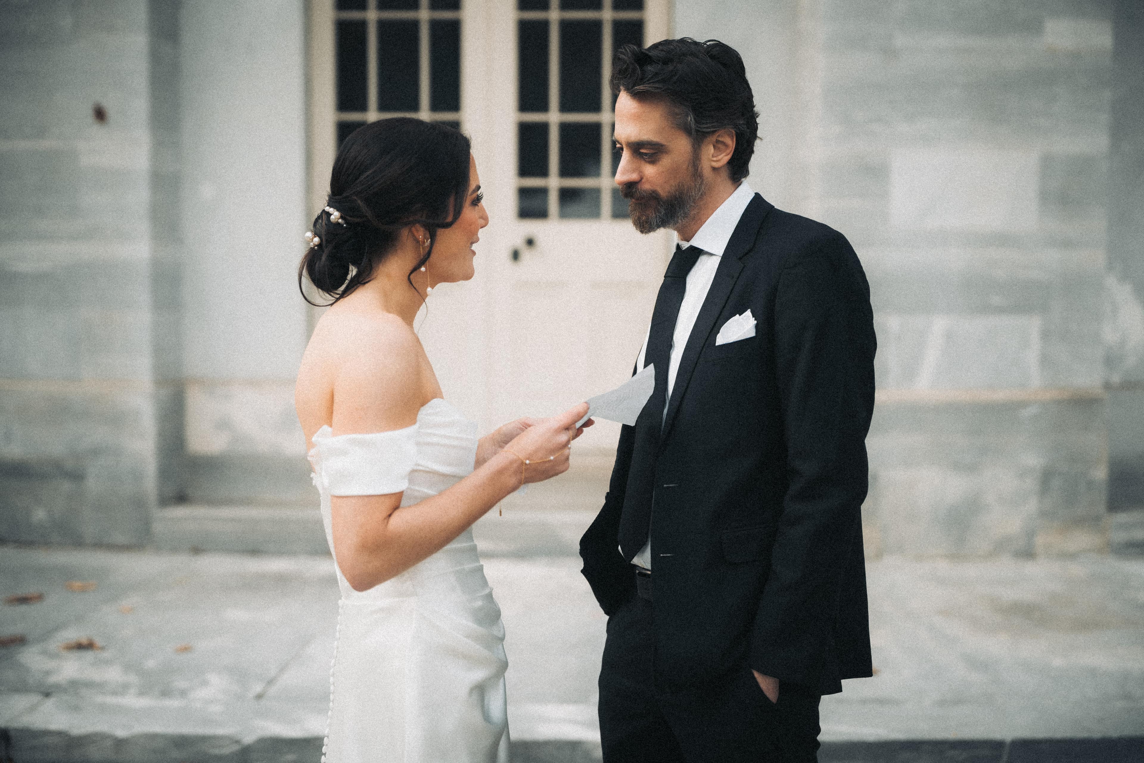 Bride reading her private vows aloud from a folded card while her partner listens, standing outside a stone building