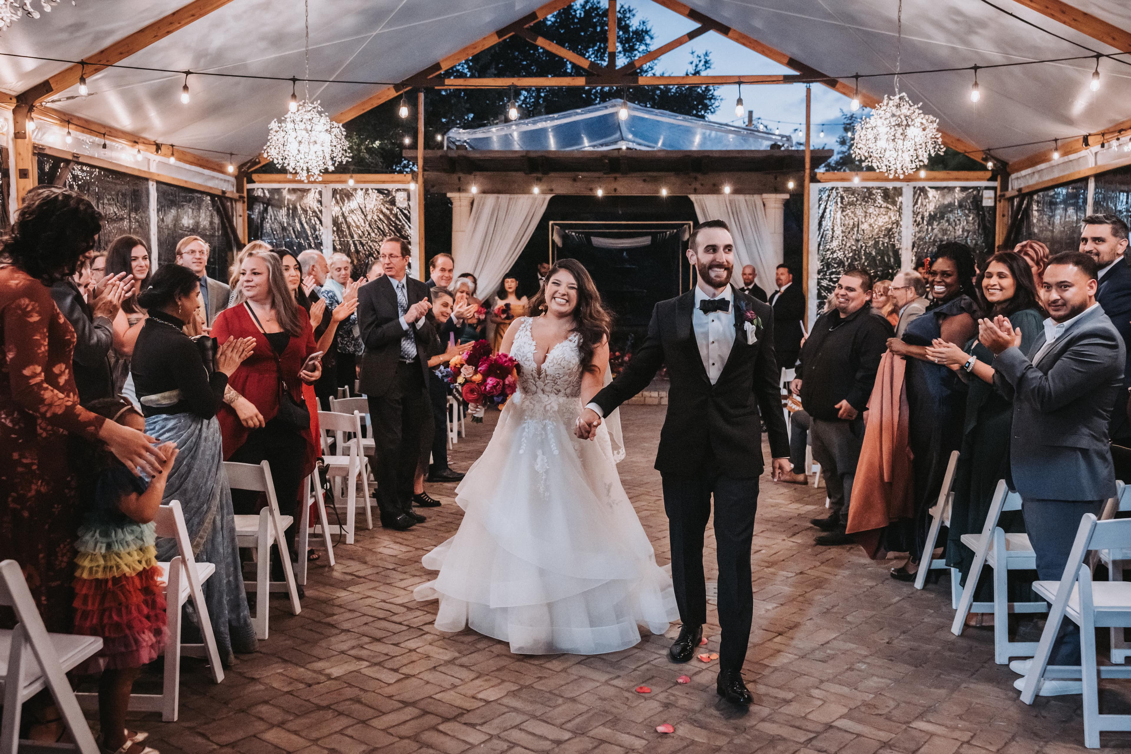 Newlyweds walking back down the aisle hand in hand as guests stand and applaud on both sides under a tented reception space