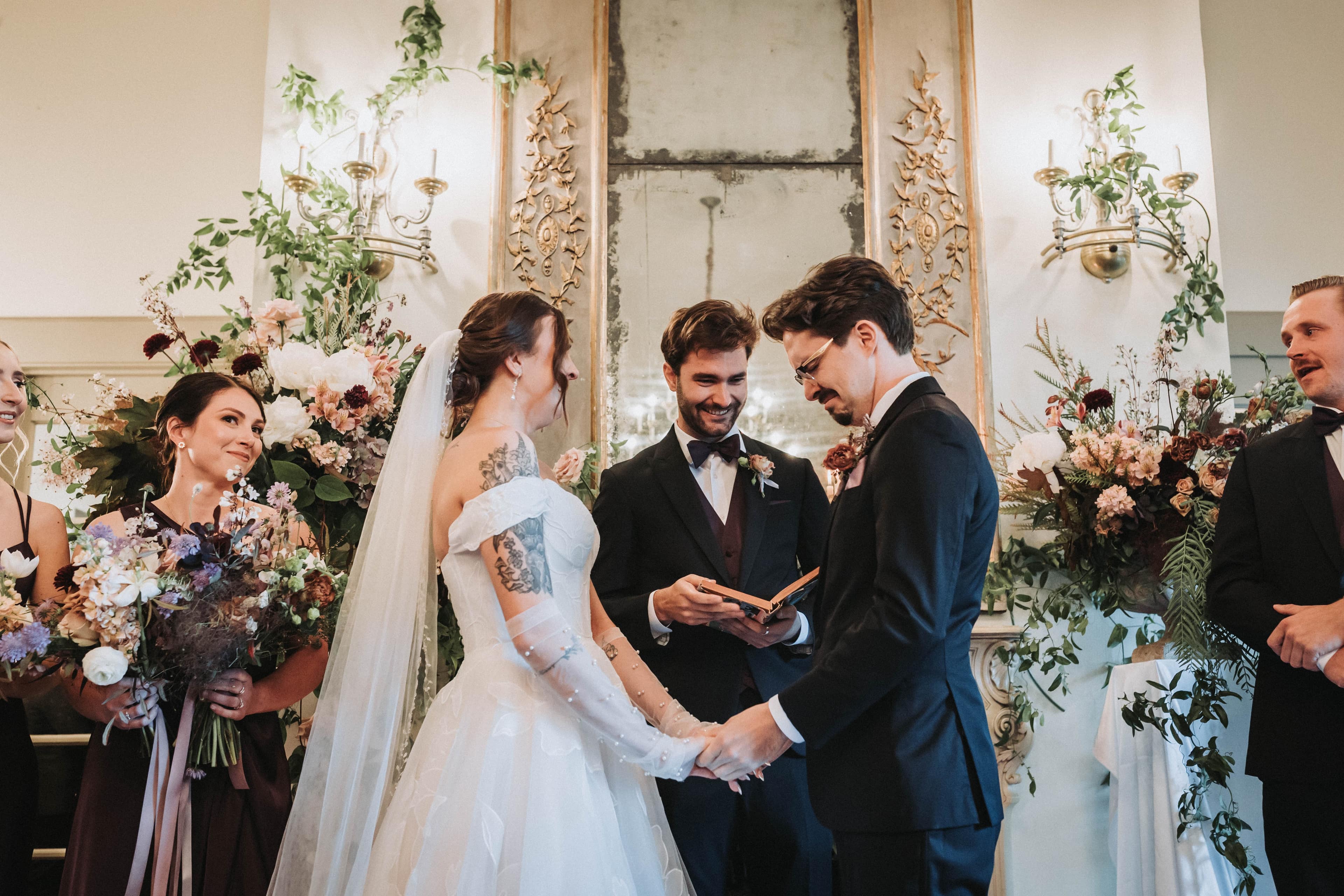 Couple holding hands through their vows in front of their wedding party and tall floral arrangements